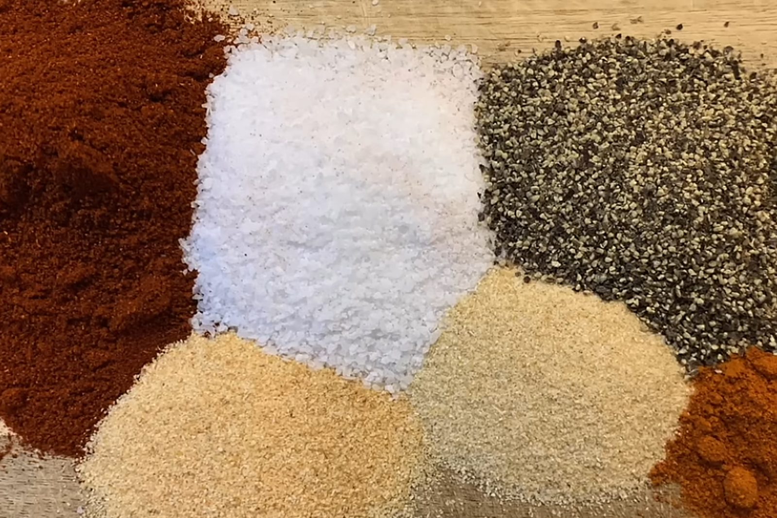 Burger rub ingredients in separate piles on a wooden board — paprika, coarse kosher salt, coarse black pepper, garlic powder, onion powder and cayenne